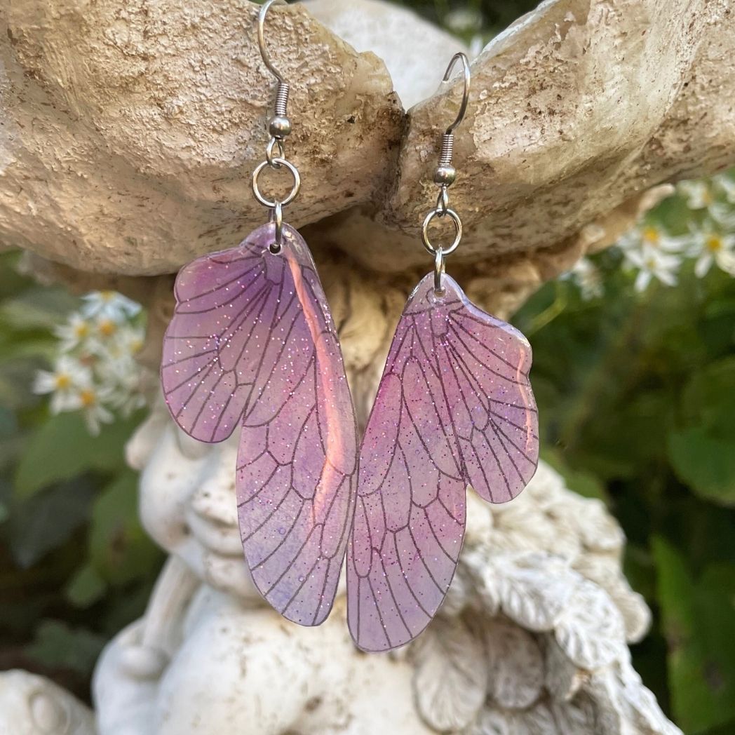 pink and purple sparkly glitter plastic fairy wing earrings hanging from a fairy garden ornament