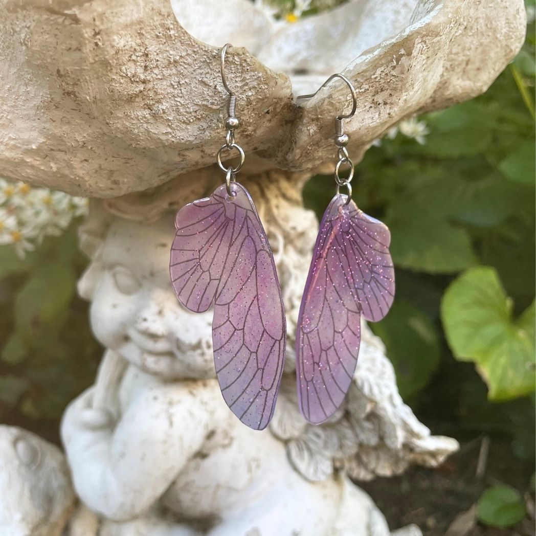 pink and purple sparkly glitter plastic fairy wing earrings hanging from a fairy garden ornament