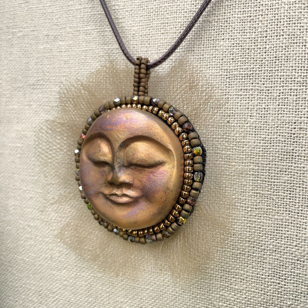 Beaded sun goddess statement pendant with sleeping face and gold beading and tulle around the edge . On a display stand 