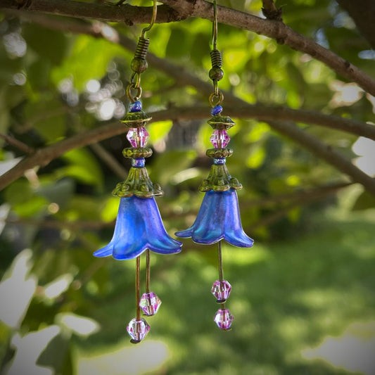 Blue lucite flower dangle earrings with purple crystals hanging on a tree branch.