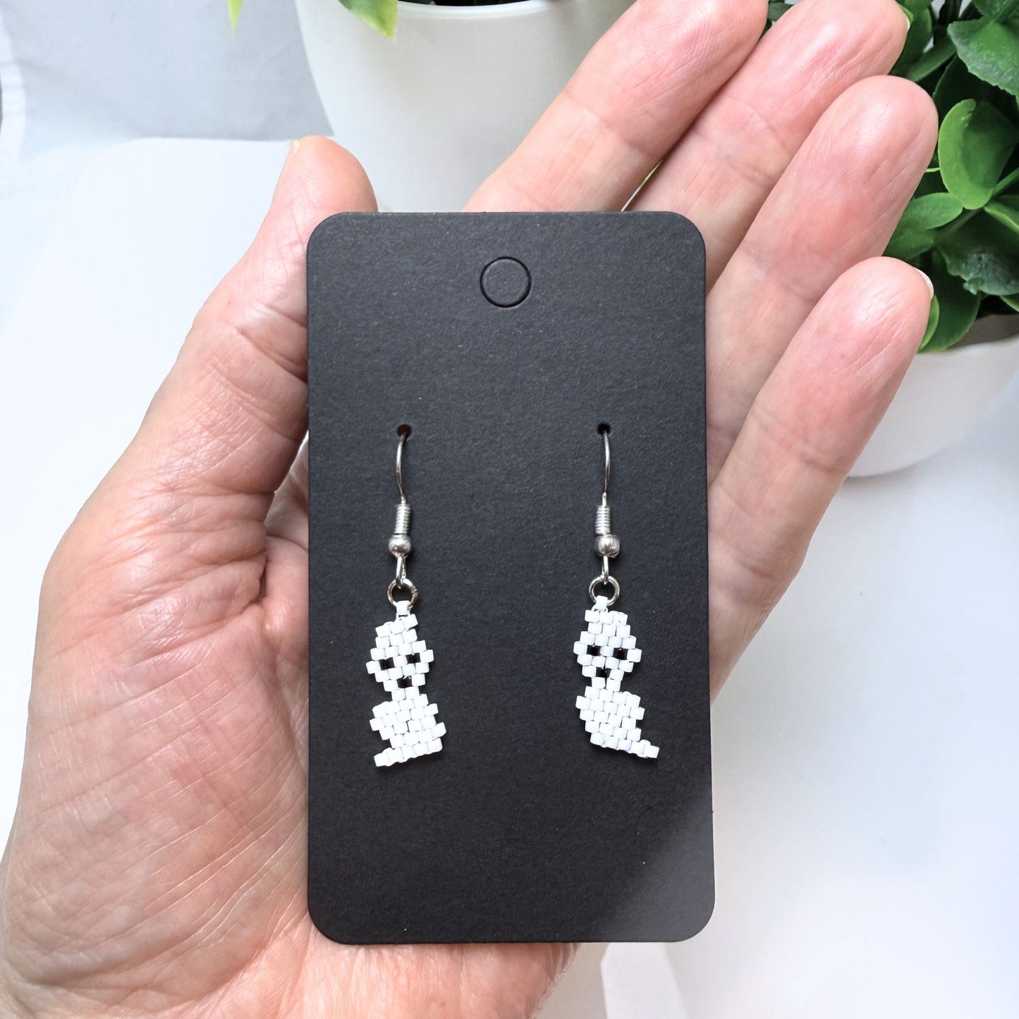 Beaded ghost earrings held in hand on black earring card showing scale