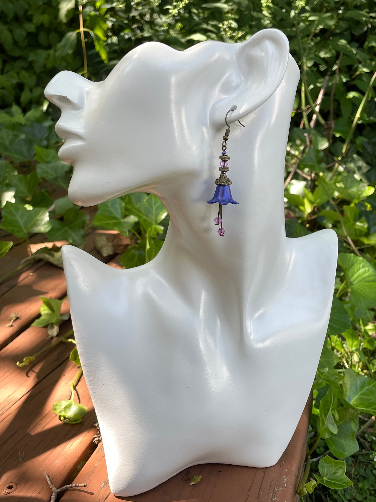 Blue flower dangle earring with antique bronze and purple beads on a model bust in a garden.