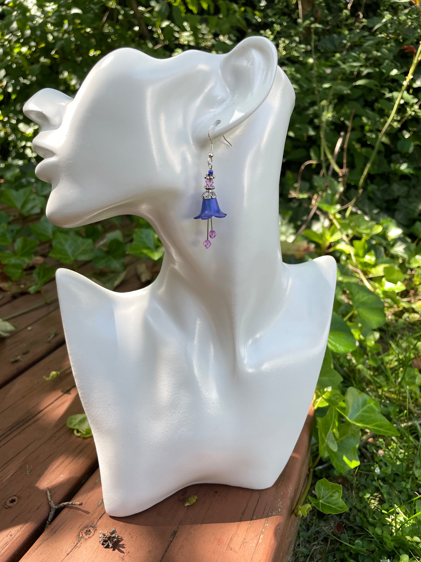 Blue flower dangle earring with silver and purple beads on a model bust in a garden.