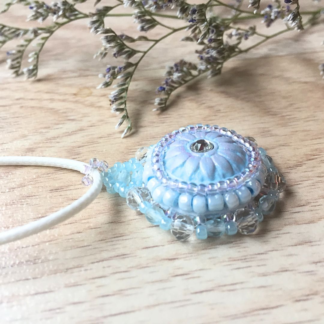 Side profile view of light blue flower necklace laying flat on a light wood surface with flower in the background.