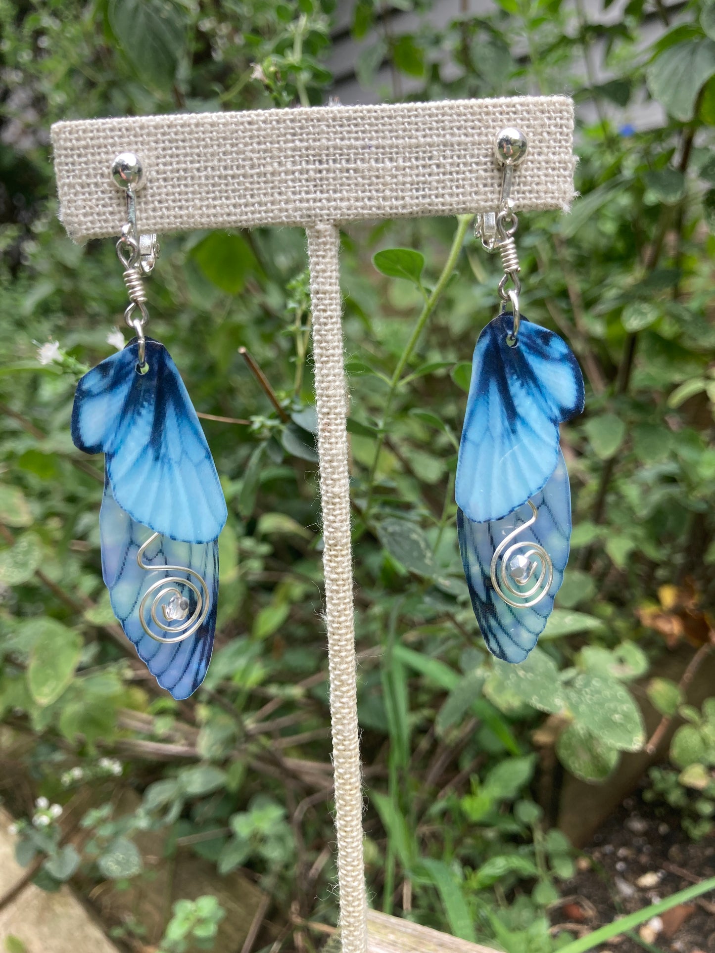 Blue butterfly clip on earrings with swirled silver wired crystals hanging from a display in a garden.