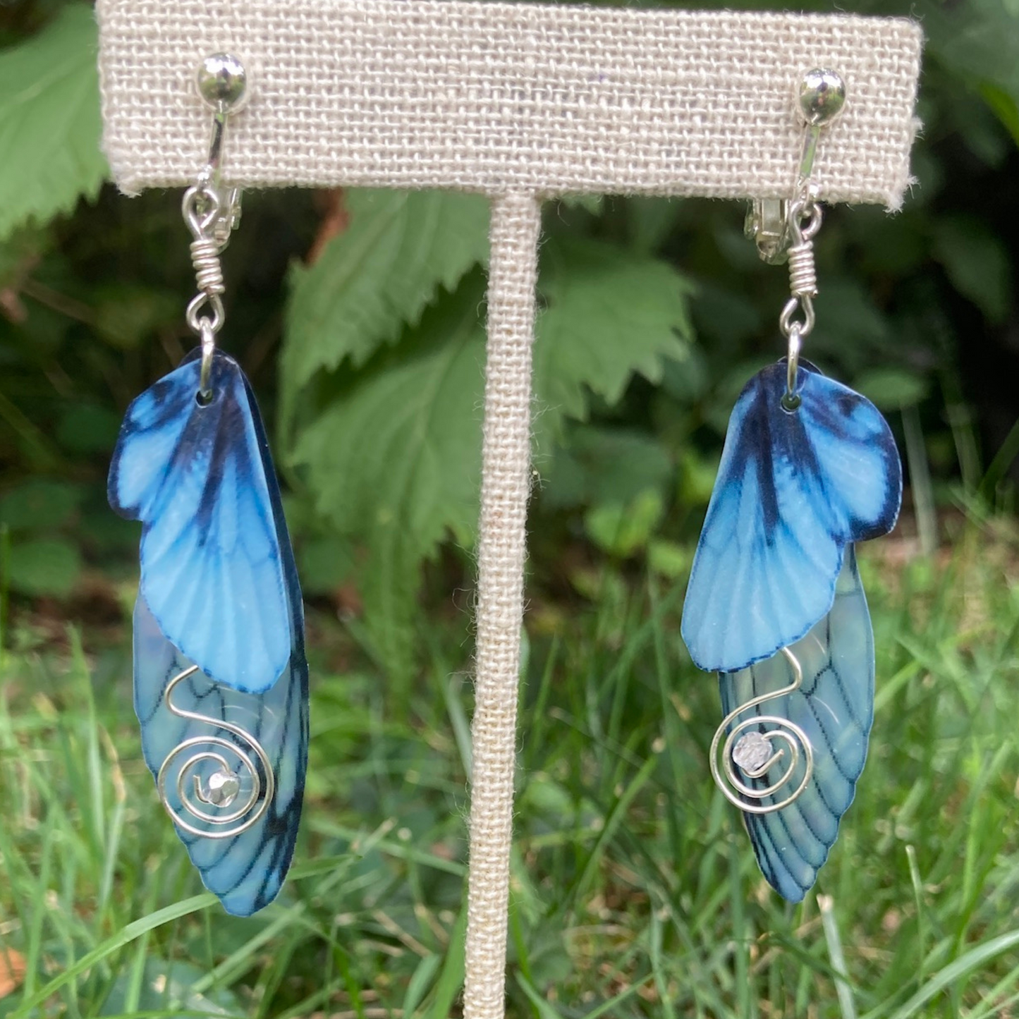 Blue butterfly clip on earrings with swirled silver wired crystals hanging from a display in a garden.