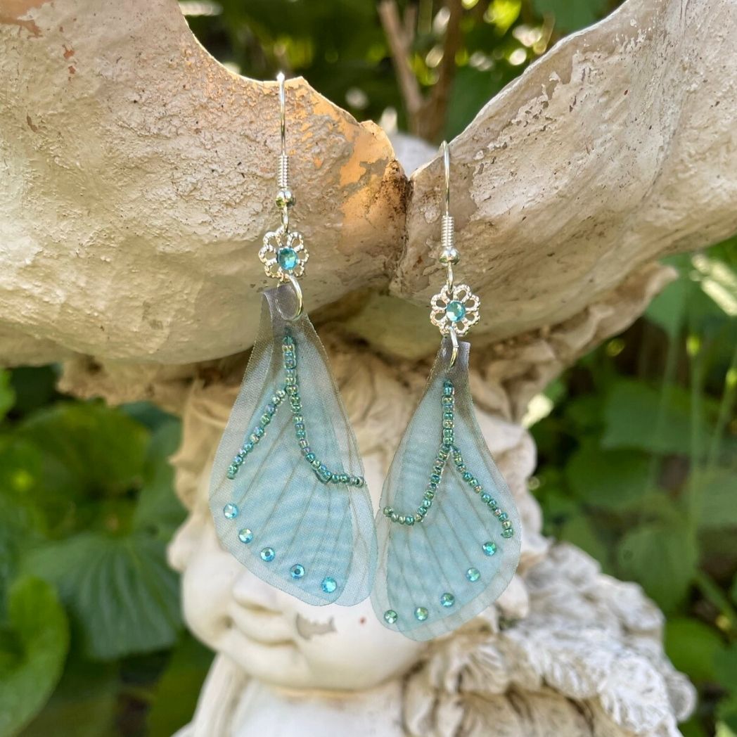 Blue chiffon butterfly wing earrings with silver crystal flower, charm and crystals on the wings hanging from a garden ornament.
