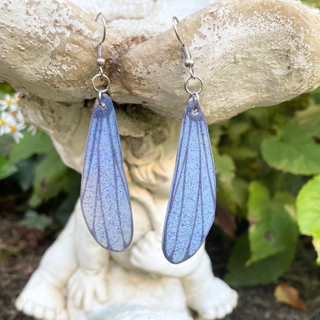 blue and gray sparkly long dangle dragonfly earrings dangling from a garden ornment.