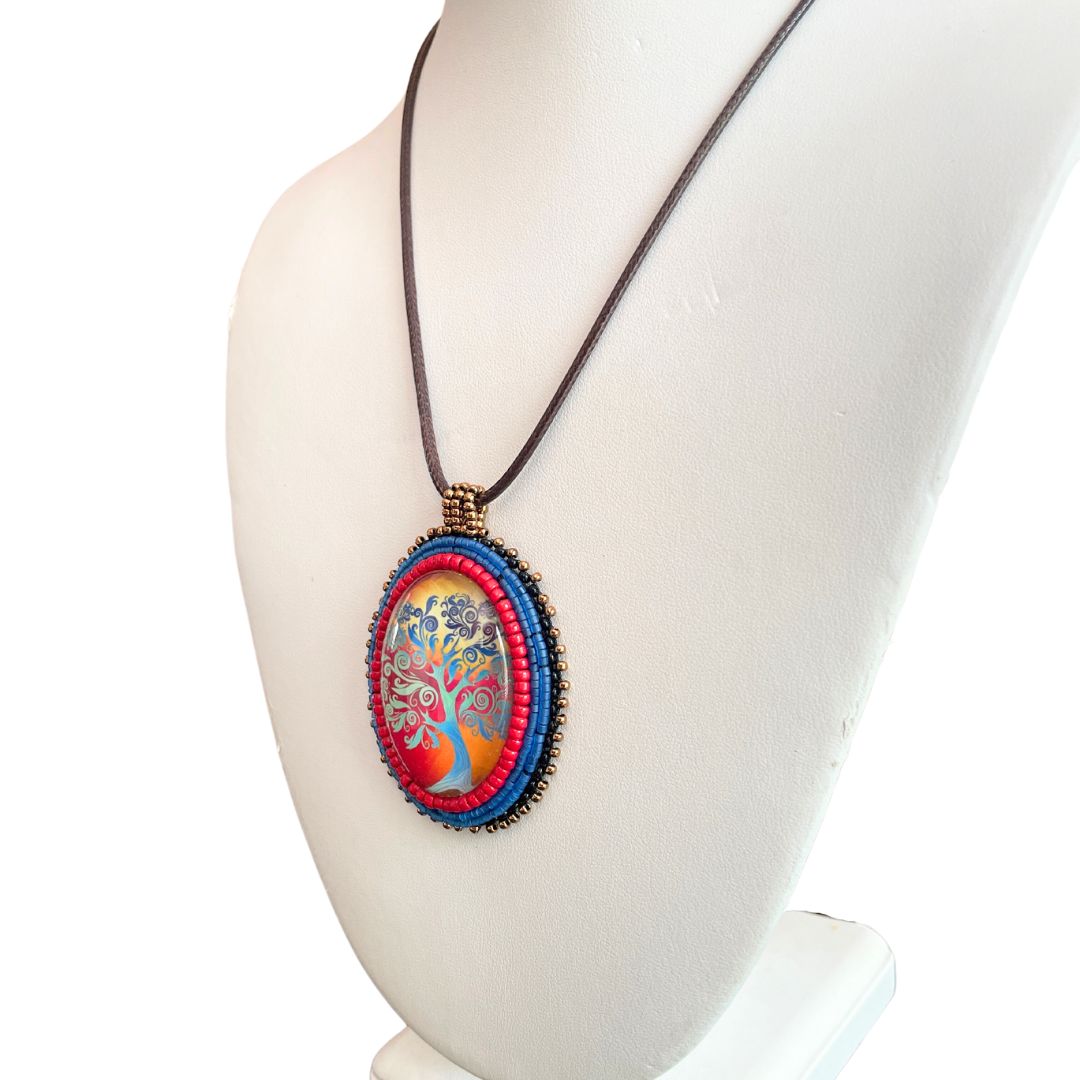 Ovsl pendant with swirly whimsical blue tree with gold and red sunset with blue, red and black glass seebead edging on a white bust