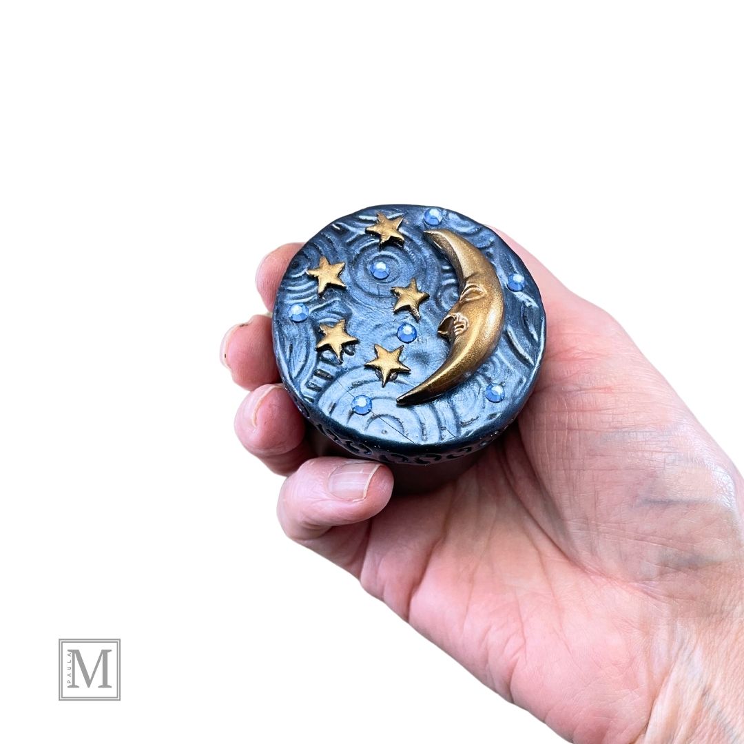 4oz candle tin with deocrative lid. Lid is blue textured polymer clay with gold crescent moon, stars an blue crystals. In model's hand.