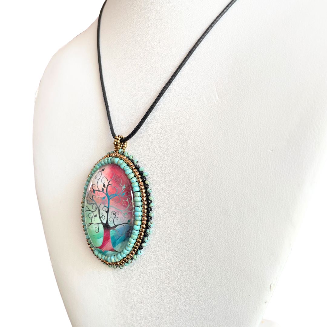 Silhoutte whimsical tree with red, blue and green background in an oval pendant with glass cabochon and green and gold seed bead edging.