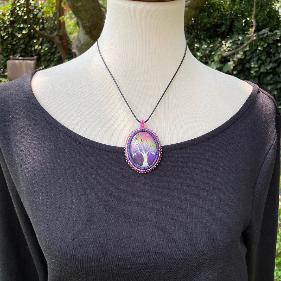 Oval pendant with whimsical tree with purple, red and pink background with pink and purple glass seed bead edging on a black cord necklace. on a mannequin.