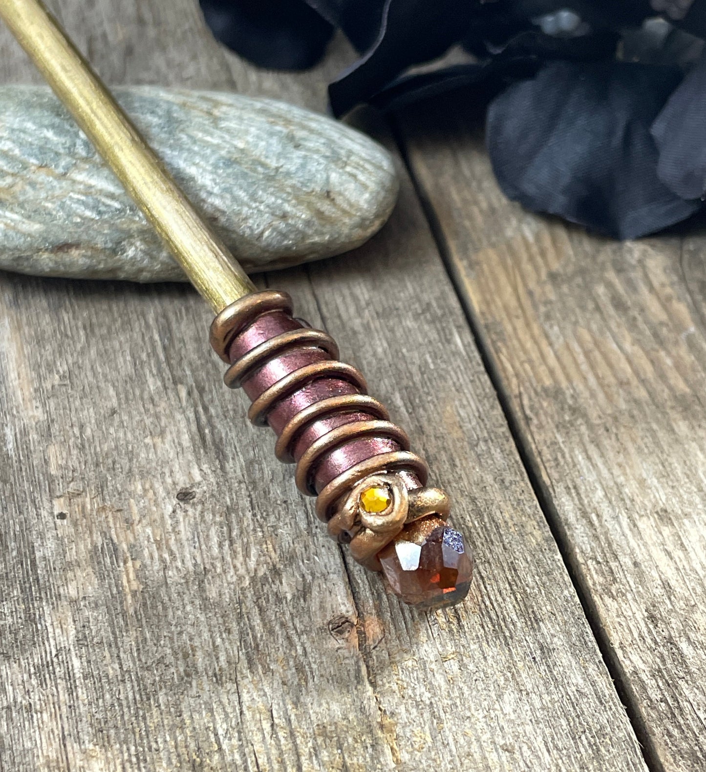 Gold painted wood hair stick with bronze and gold spiral clay hand painted details with dark AmberAB crystal. Resting on a pebble with a black flower to one side.