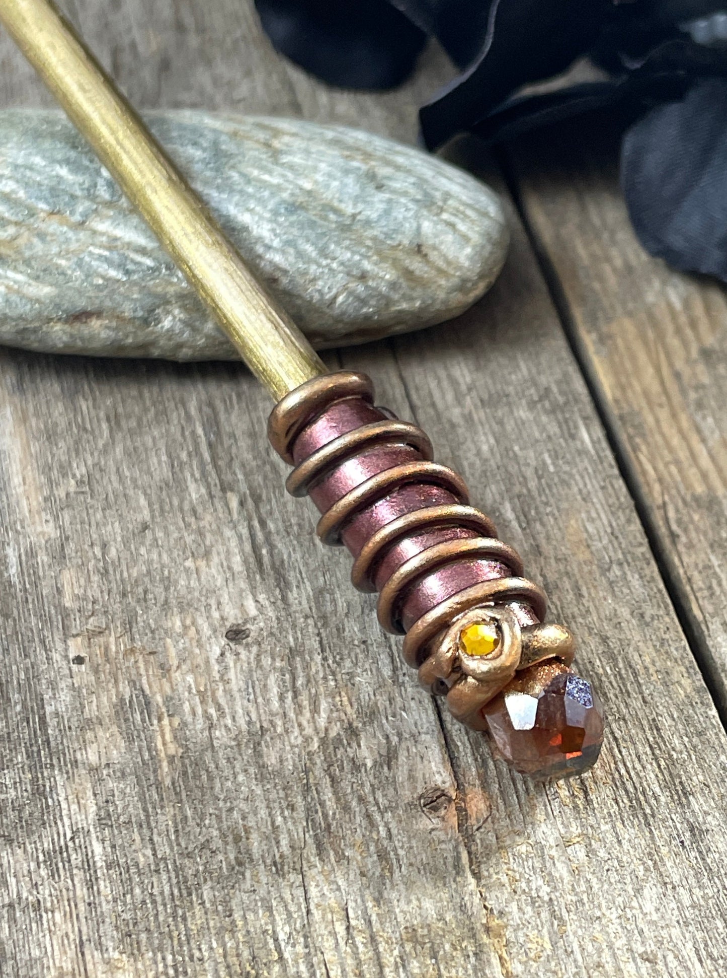 Gold painted wood hair stick with bronze and gold spiral clay hand painted details with dark AmberAB crystal. Resting on a pebble with a black flower to one side.