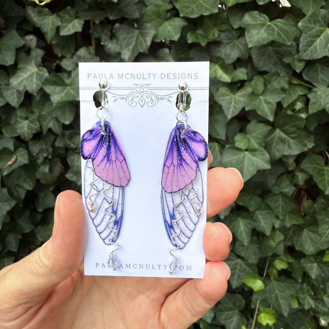 Close-up of handmade purple fairy wing clip-on earrings in silver finish with glittered wings and crystals.