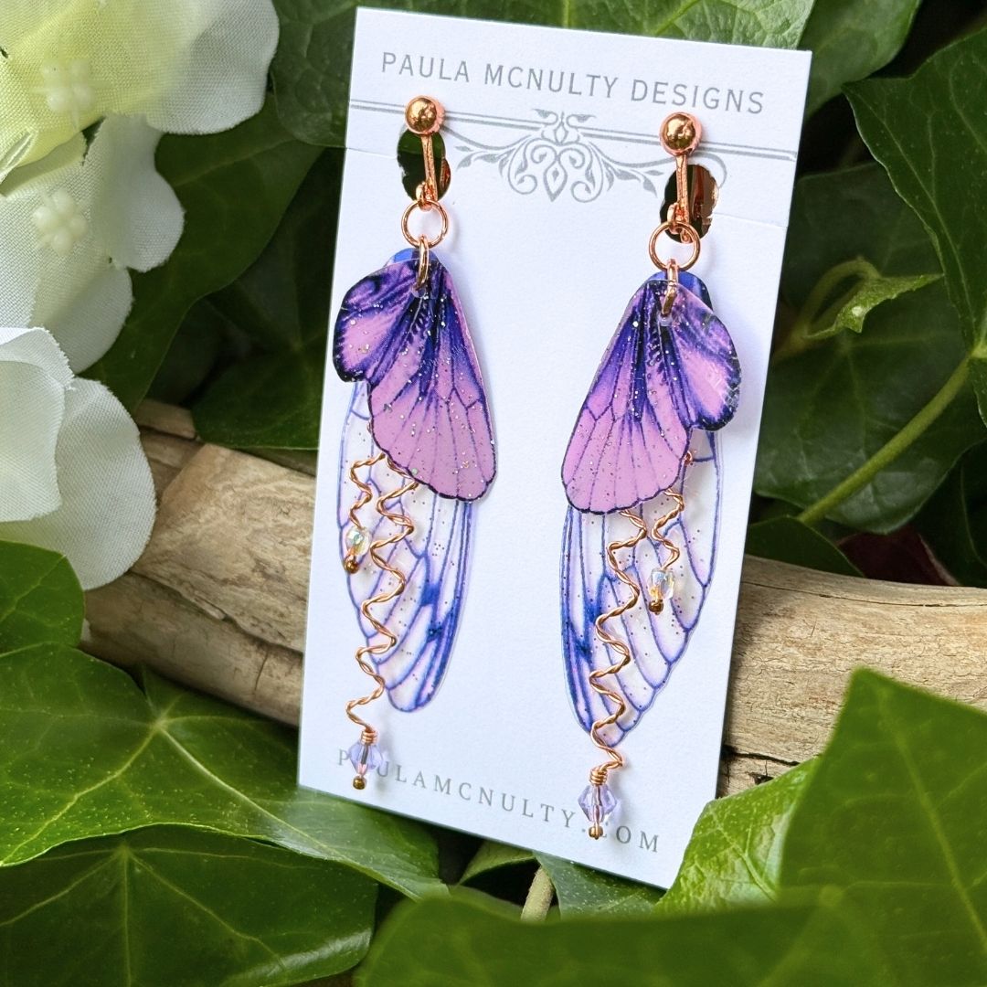 Purple fairy wing earrings in copper finish displayed among ivy leaves, handmade fantasy jewelry gift.