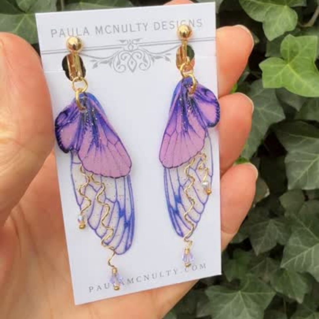 Hand holding purple clip-on butterfly wing earrings with gold finish and comfort pads by Paula McNulty Designs.