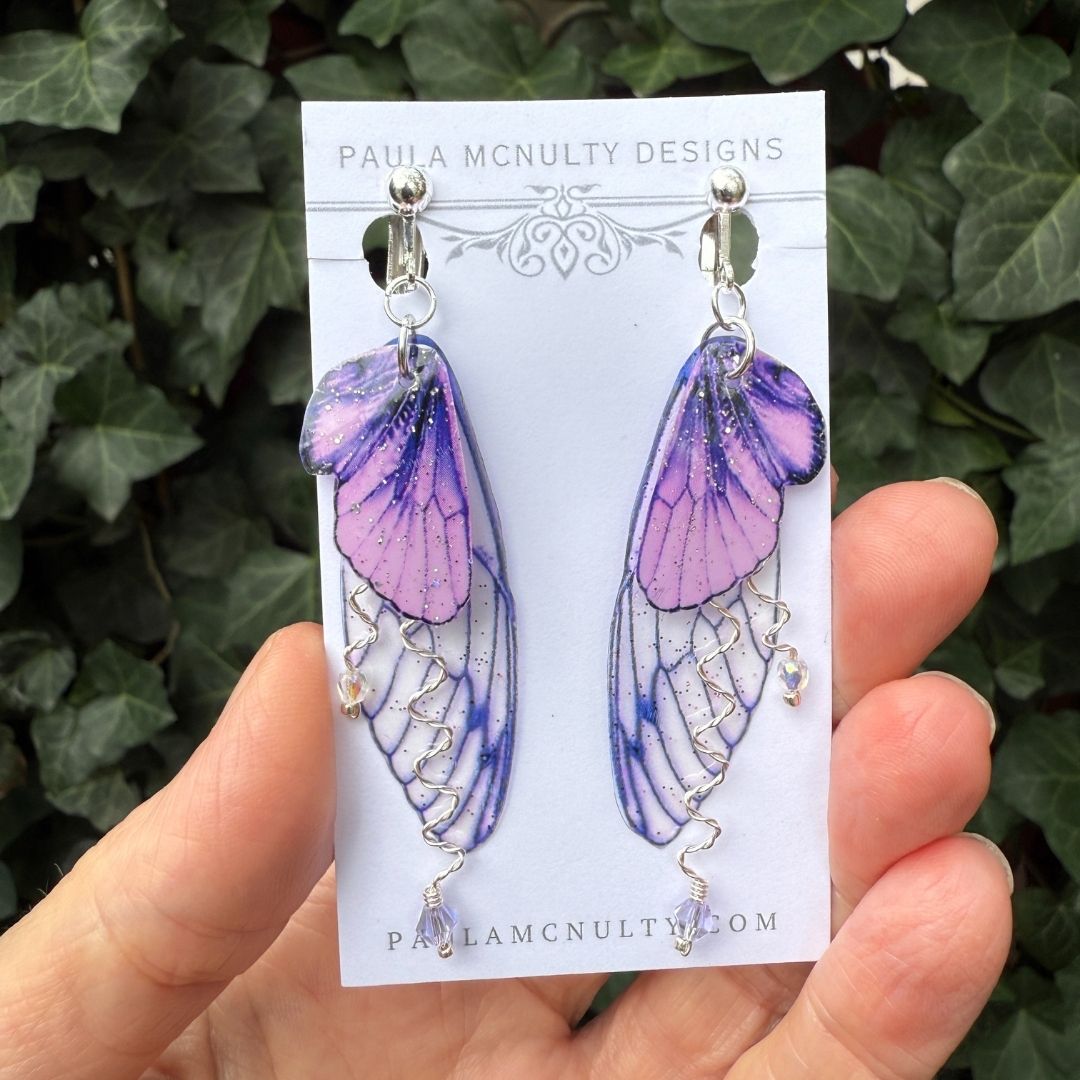Close-up of handmade purple fairy wing clip-on earrings in silver finish with glittered wings and crystals.