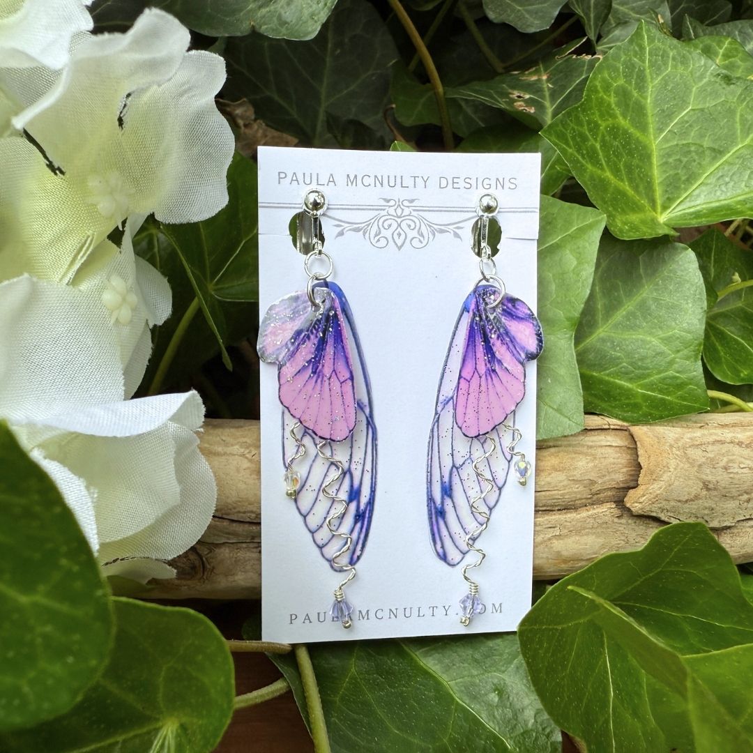 Close-up of handmade purple fairy wing clip-on earrings in silver finish with glittered wings and crystals.