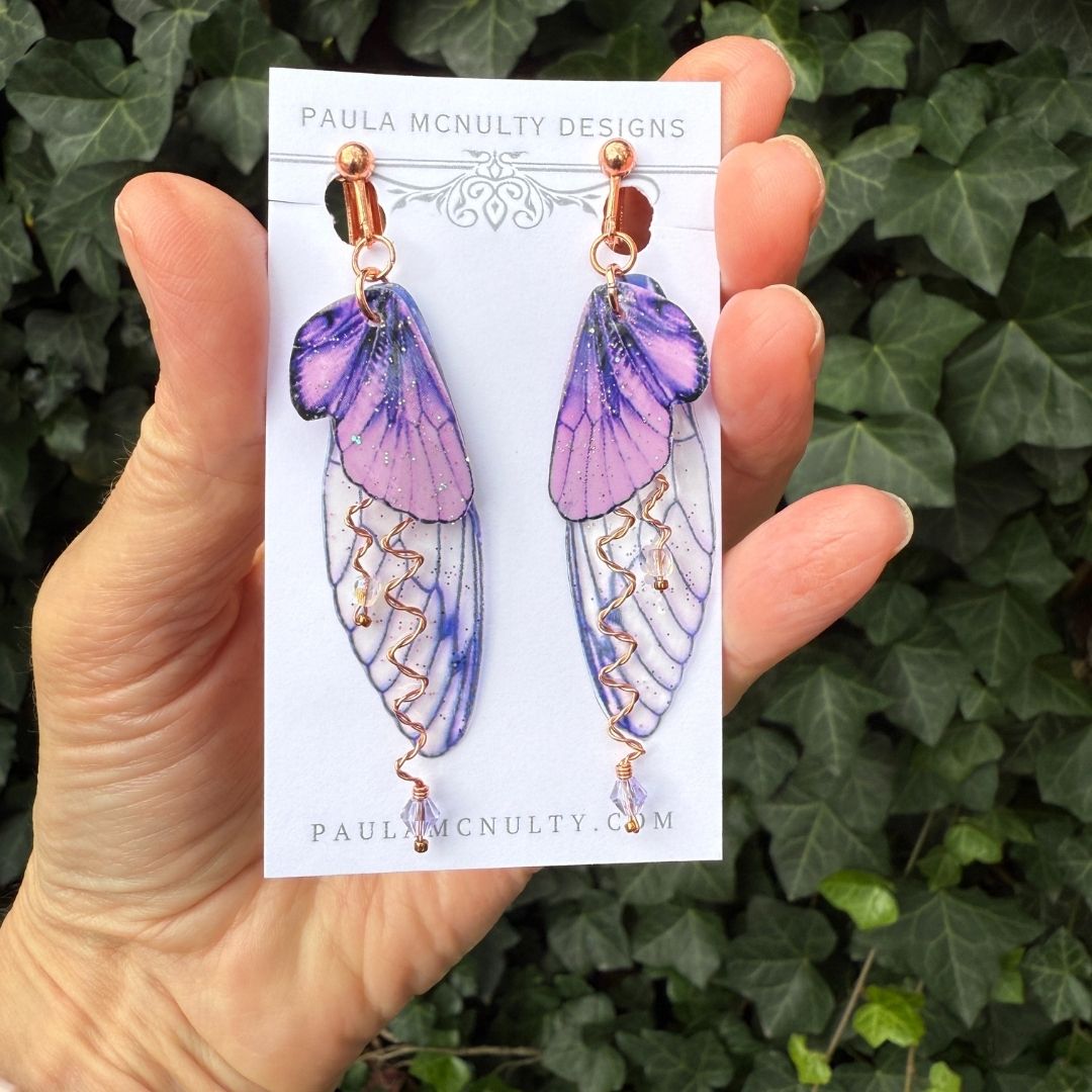 Close-up of handmade purple fairy wing clip-on earrings in copper finish with glittered wings and crystals.