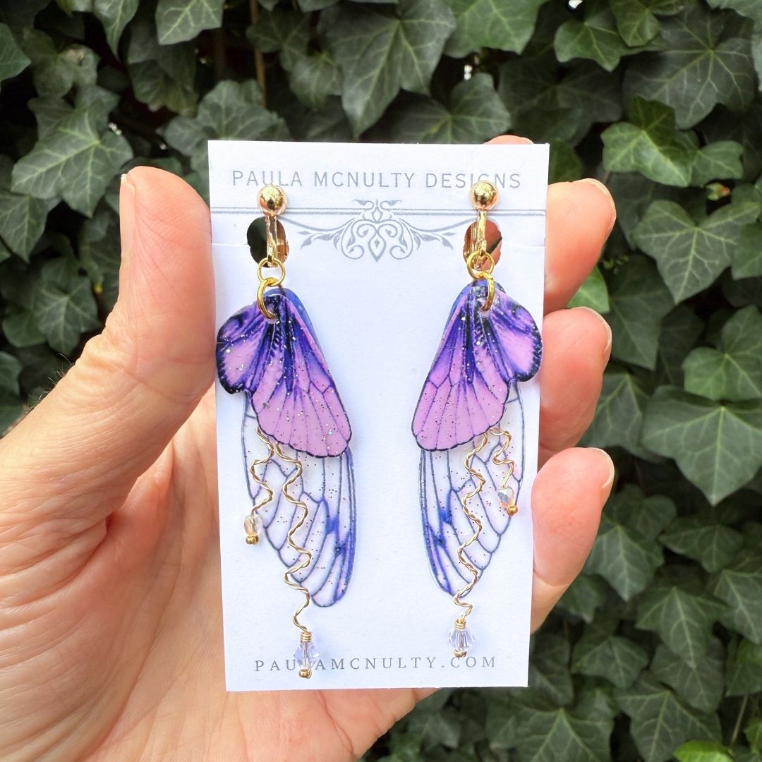 Close-up of handmade purple fairy wing clip-on earrings in gold finish with glittered wings and crystals.