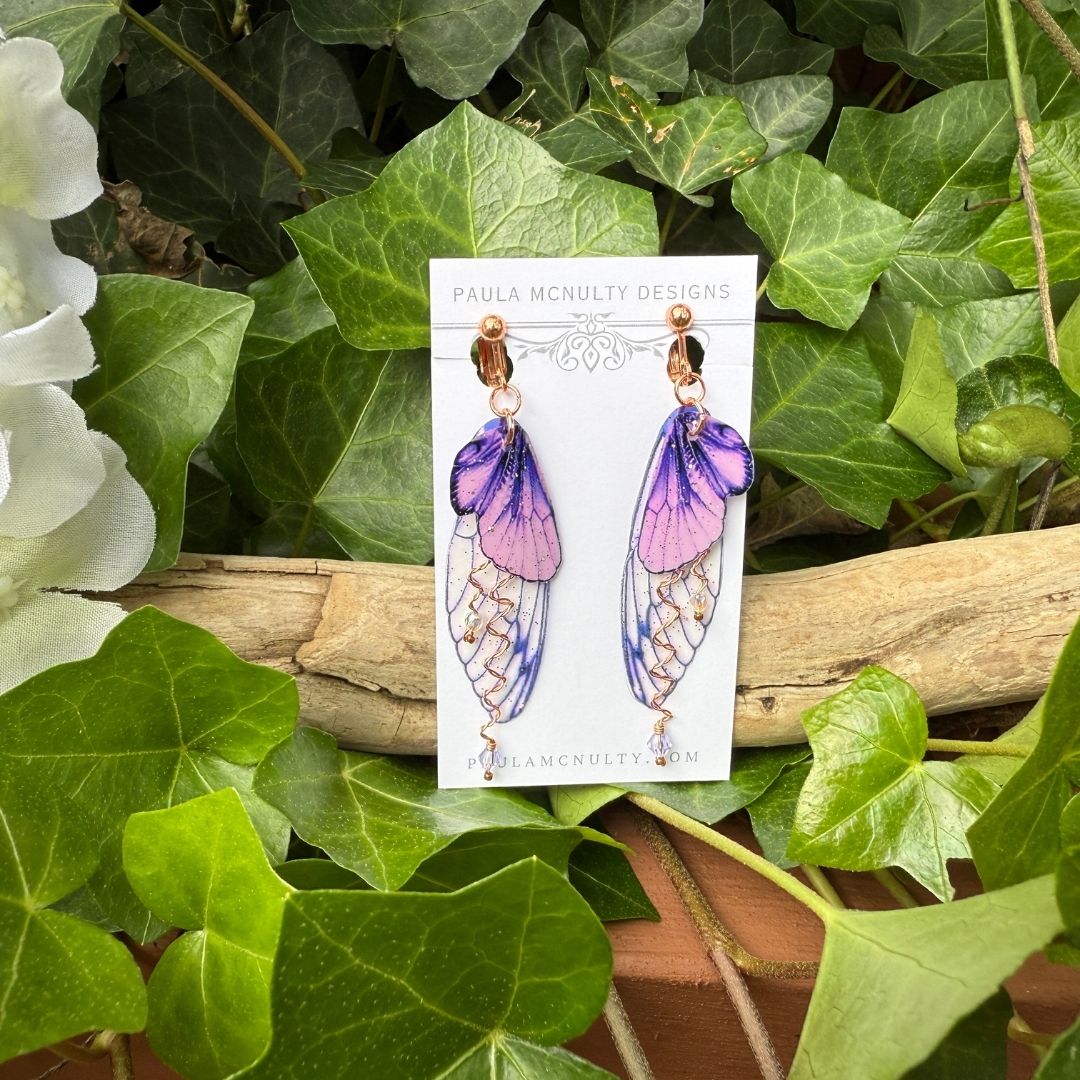 Close-up of handmade purple fairy wing clip-on earrings in silver finish with glittered wings and crystals.