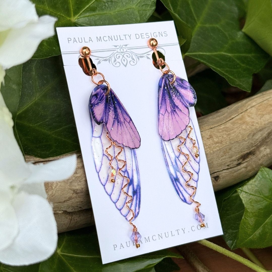 Purple fairy wing earrings in copper finish displayed among ivy leaves, handmade fantasy jewelry gift.
