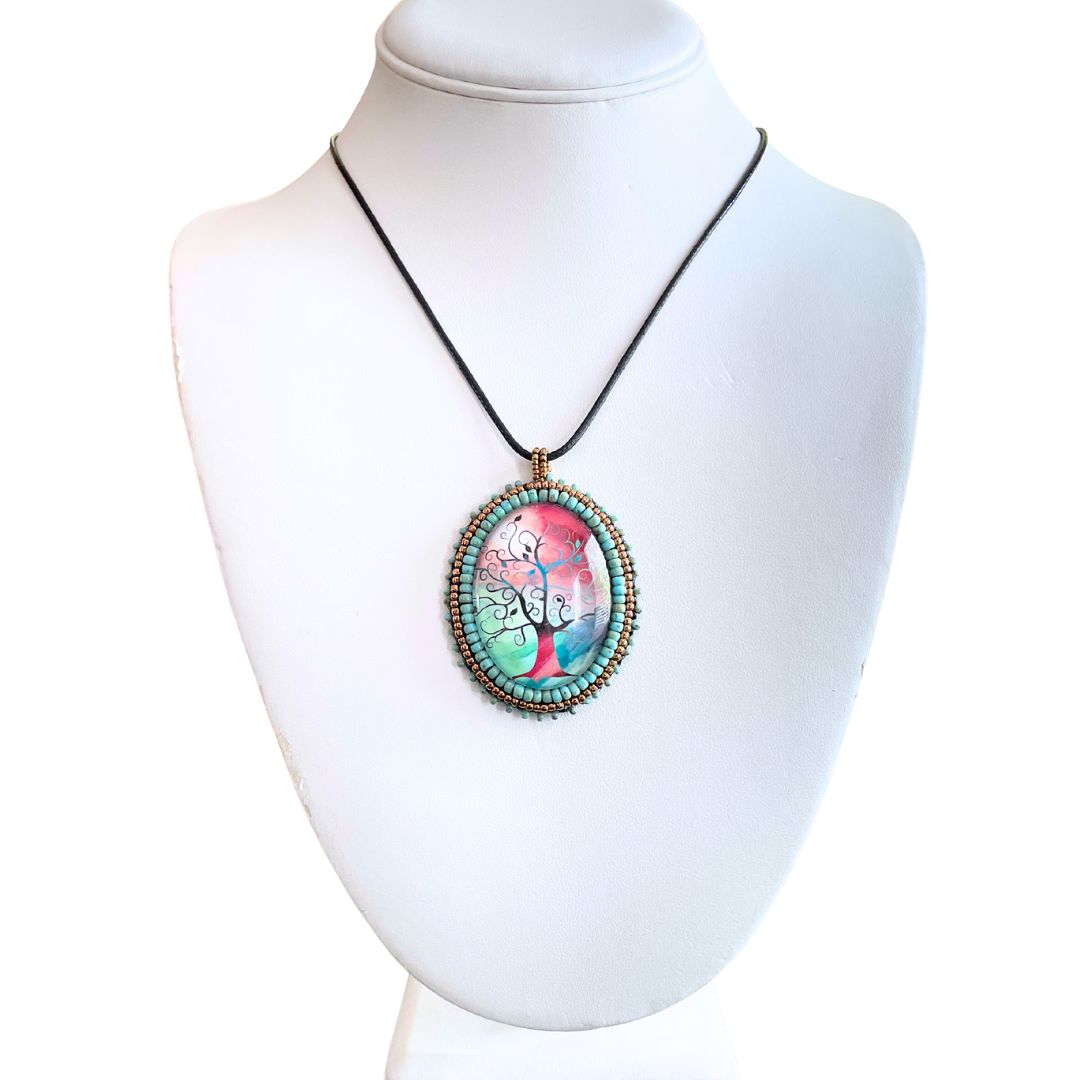 Silhoutte whimsical tree with red, blue and green background in an oval pendant with glass cabochon and green and gold seed bead edging on a bust,