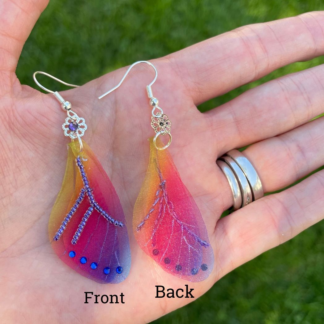 Colorful rainbow sheer fabric with beading and blue crystals. A flower bead with a crystal connects the wings to the ear wires. Display in a hand.