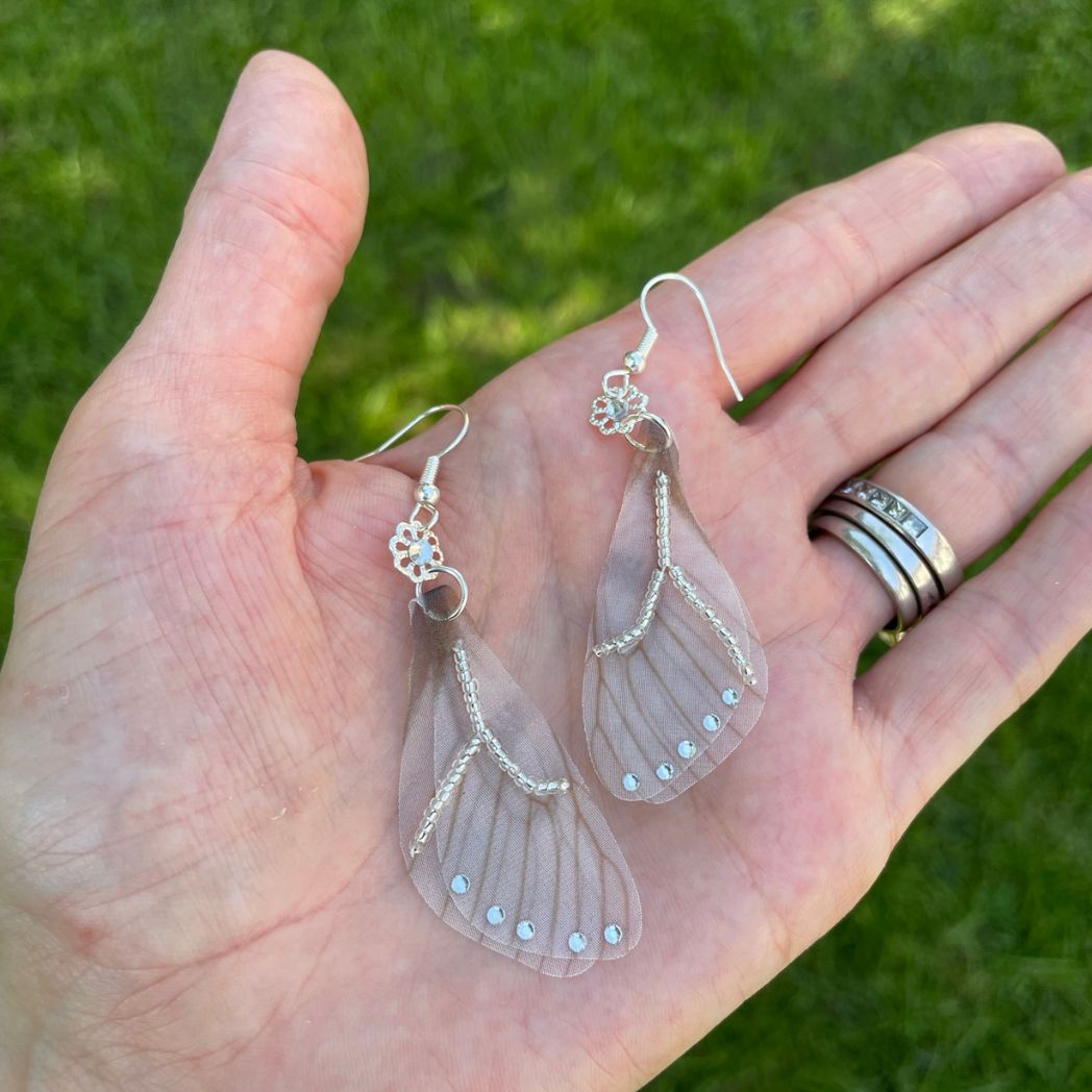 Silver beaded gray sheer fabric butterfly wing earrings with crystals and a flower bead connecting to the ear wires in a hand