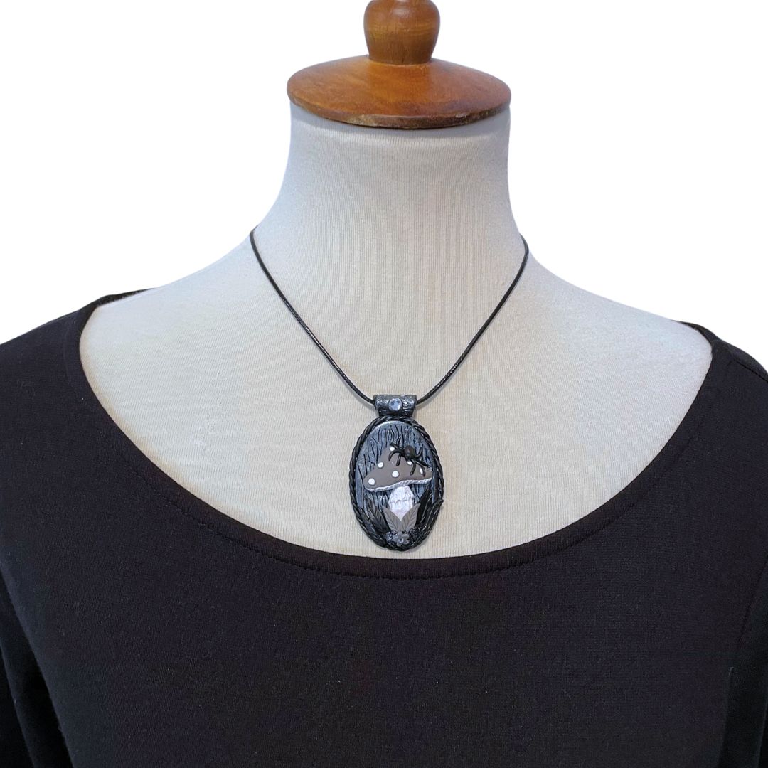 A model bust wearing an oval pendant in silver and black with a spider sitting on a gray mushroom with flower detail at the base of the mushroom.