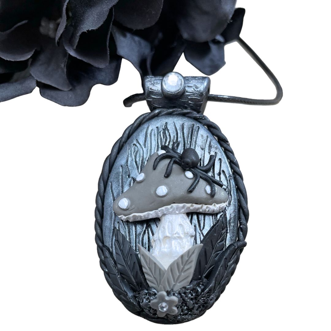 oval pendant in silver and black with a spider sitting on a gray mushroom with flower detail at the base of the mushroom. A black flower is in the top left corner.