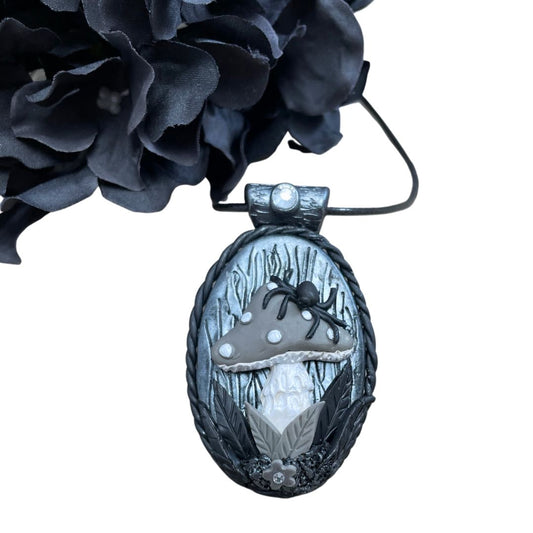 oval pendant in silver and black with a spider sitting on a gray mushroom with flower detail at the base of the mushroom. A black flower is in the top left corner.