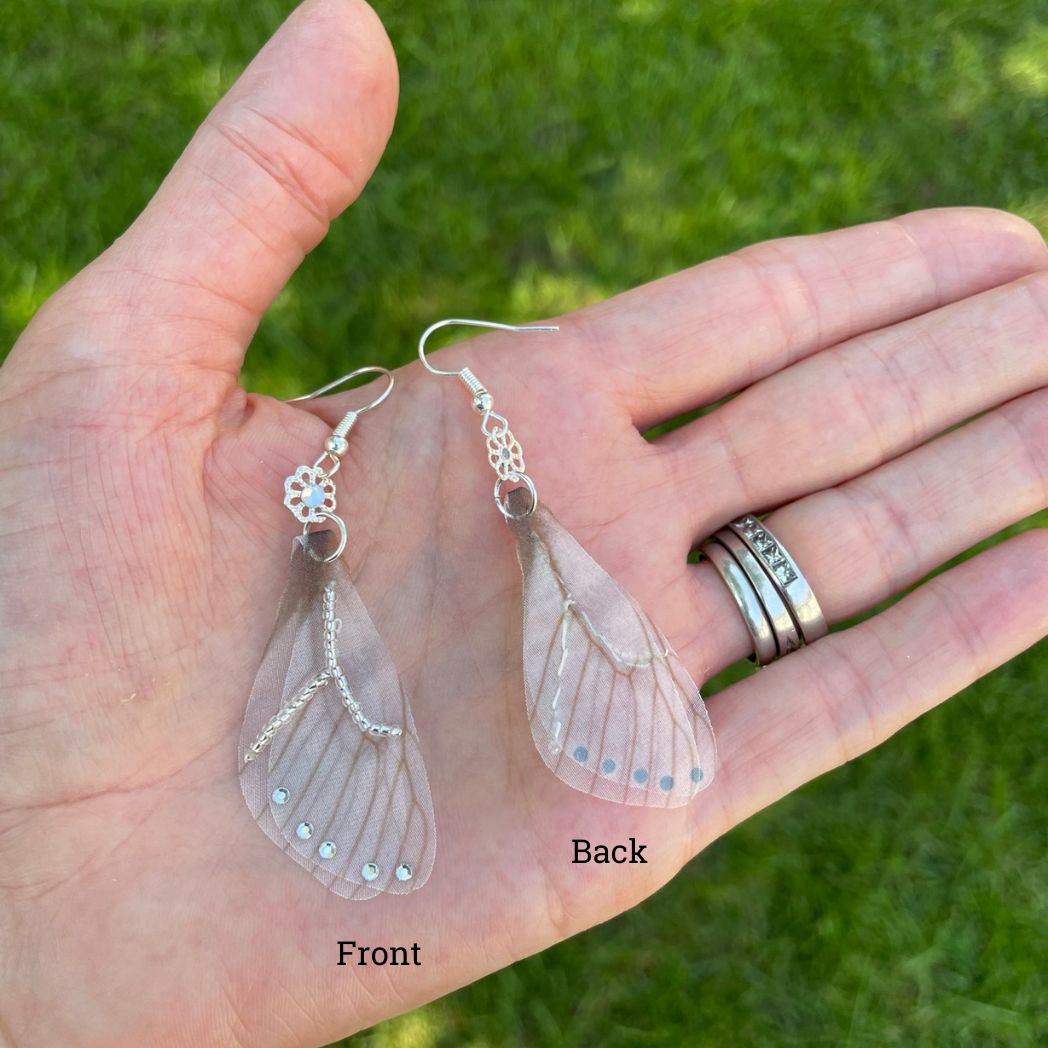 Silver beaded gray sheer fabric butterfly wing earrings with crystals and a flower bead connecting to the ear wires in a hand.