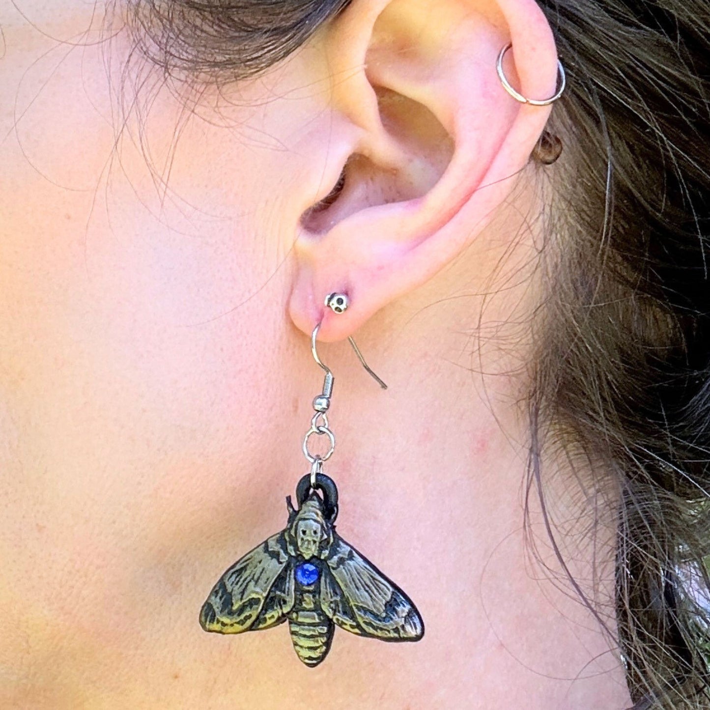 A close-up of a person's ear wearing a dangling moth-shaped earring. The earring has a silver fishhook wire, a small silver bead, and a black top with a yellow-and-black moth body and wings, plus a small blue gemstone accent.