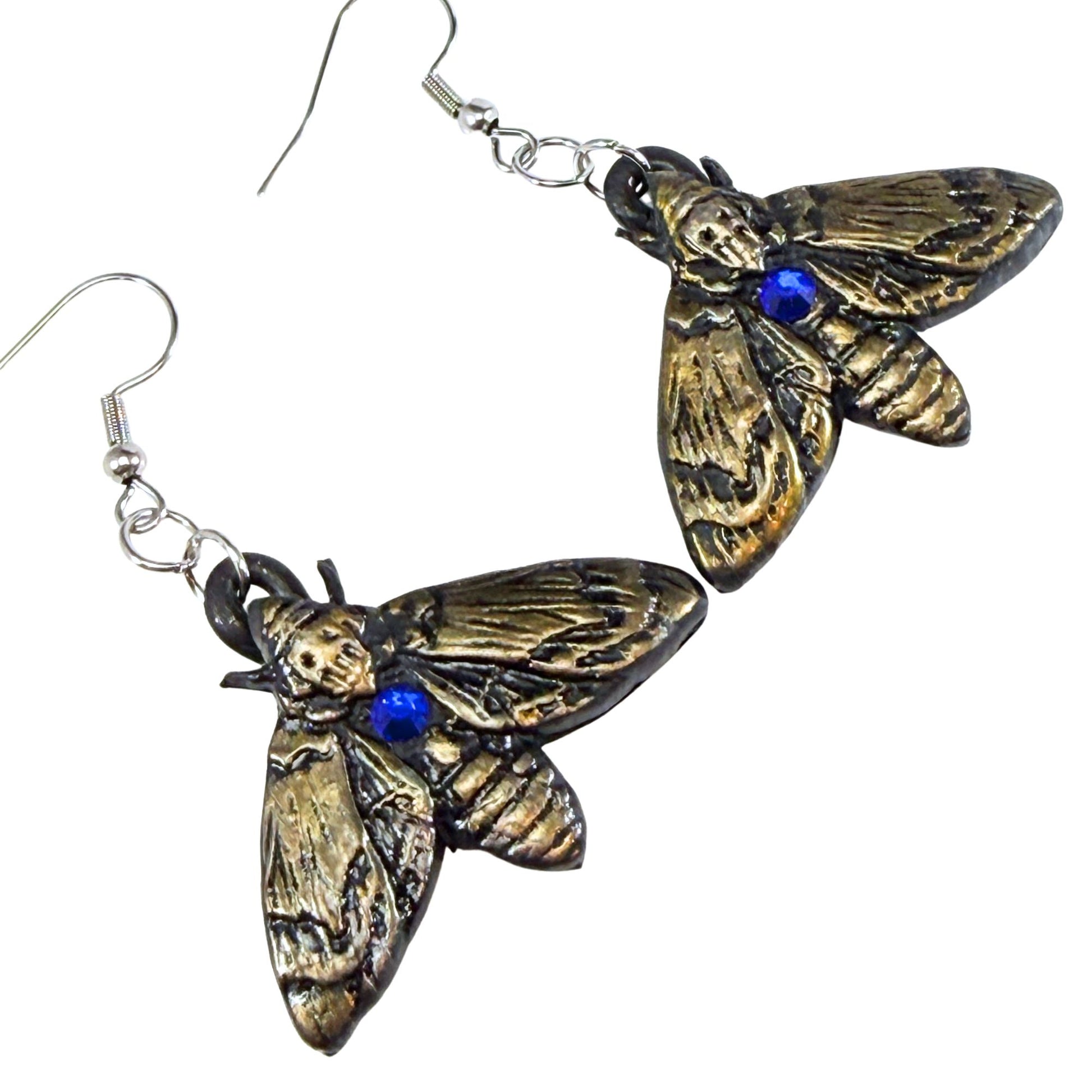 A pair of gothic moth drop earrings with textured bronze wings and a small blue gemstone accent on the thorax, suspended from silver fishhook wires against a plain white background.
