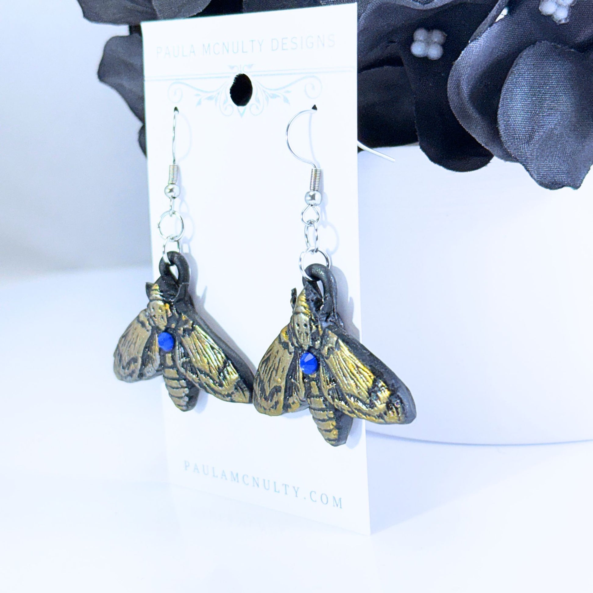 A pair of dangling moth-shaped earrings with antiqued gold and black wings, each accented by a small blue bead at the center, displayed on a white card labeled Paula McNulty Designs.