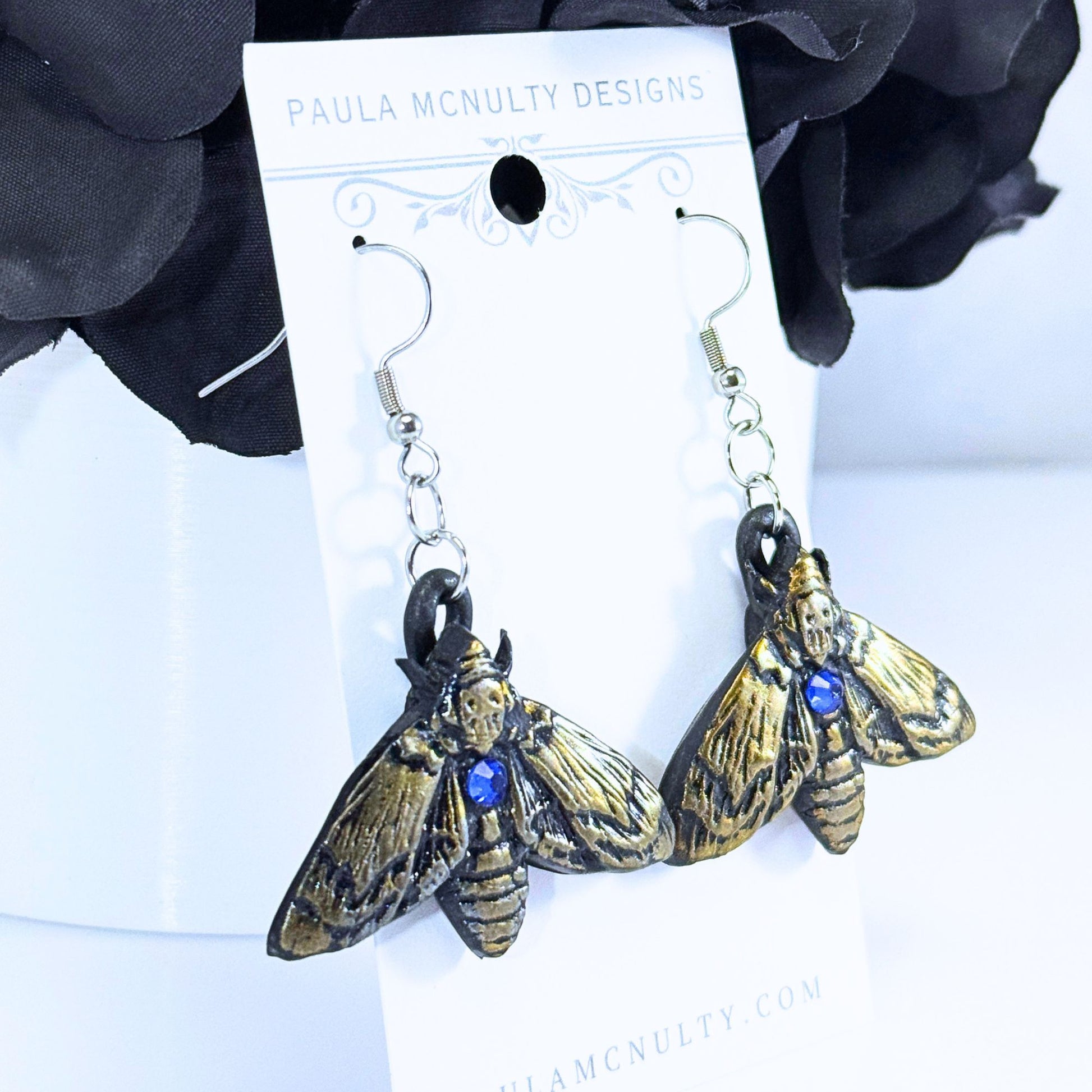 A pair of Paula McNulty Designs moth earrings with gold-toned wings, dark bodies, and small blue gemstone accents, hanging from silver fishhook wires on a white display card.