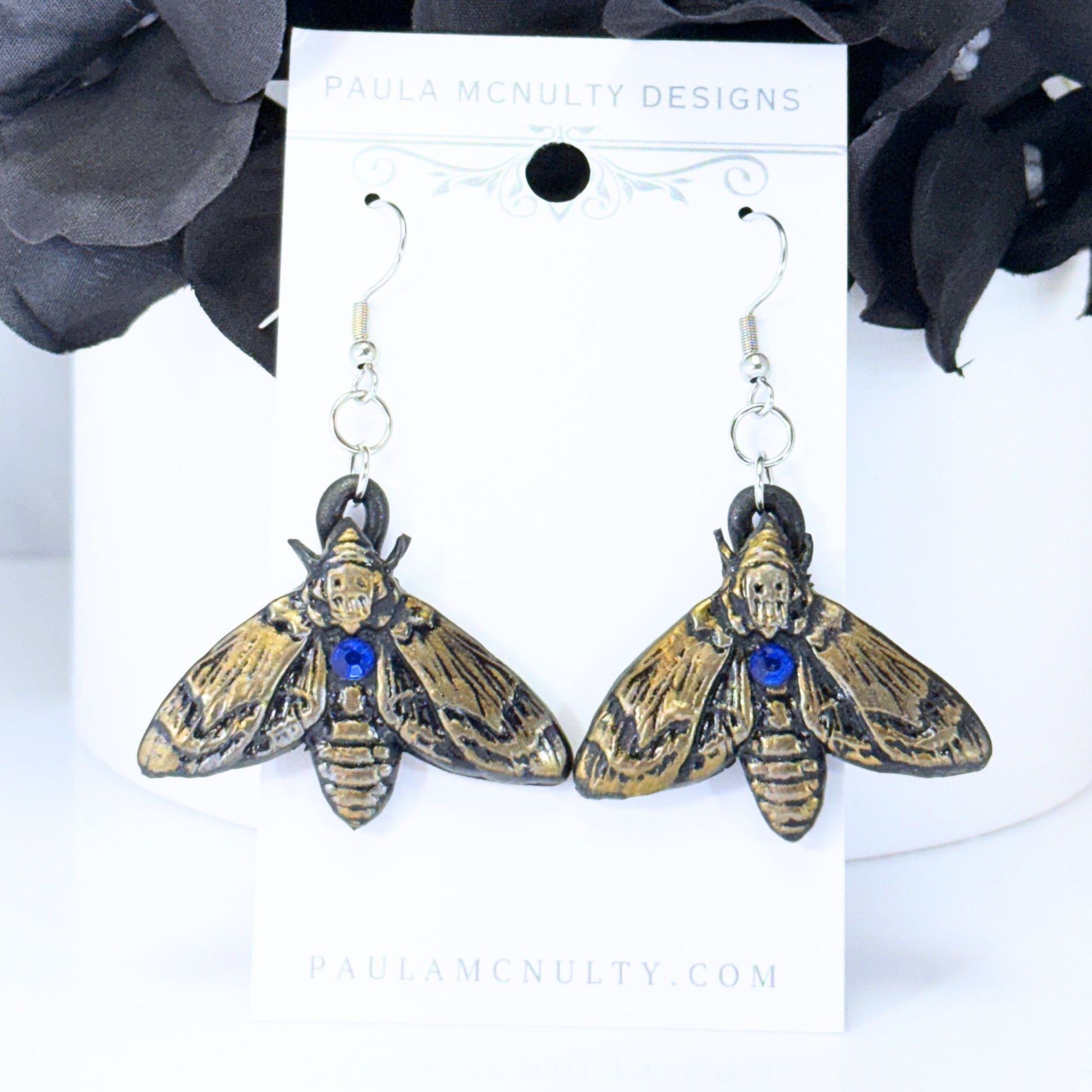 A pair of gothic moth drop earrings by Paula McNulty Designs, featuring antique bronze-toned wings with black detailing and a small blue crystal accent on the thorax, displayed on a white brand card.