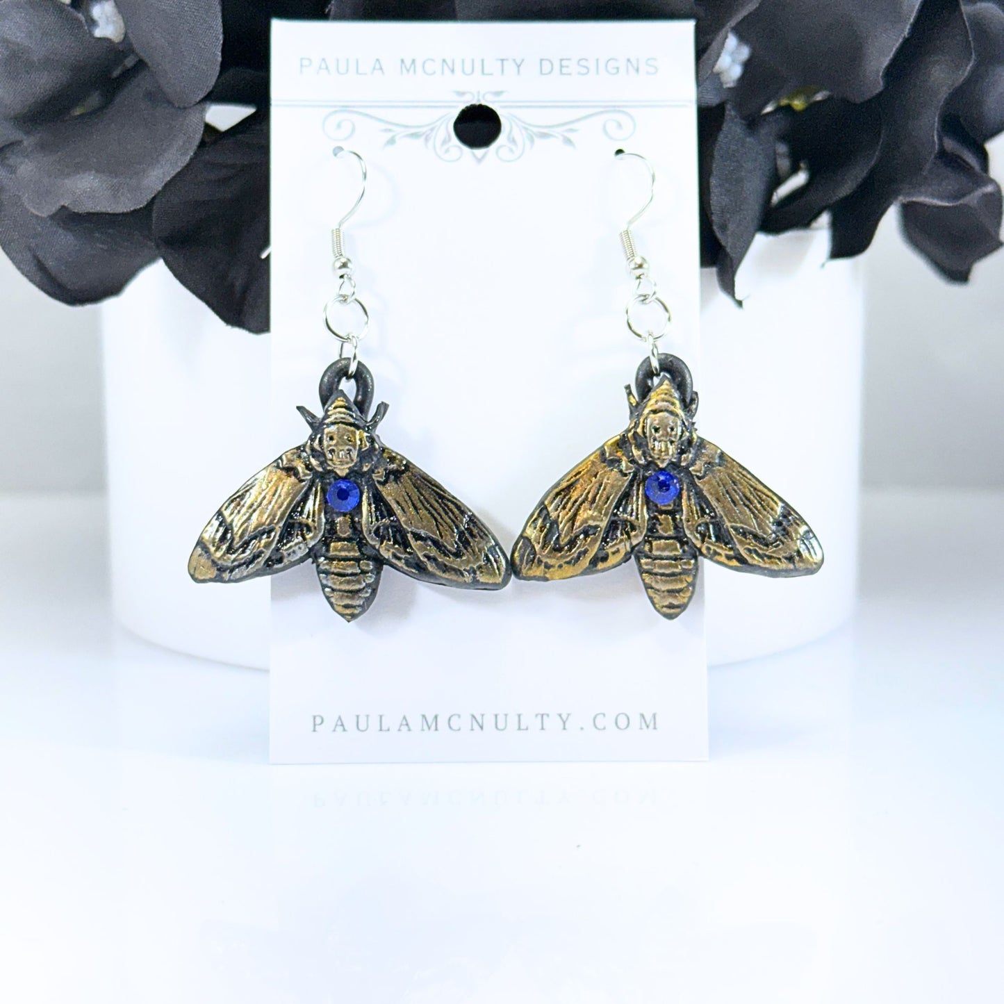 A pair of moth-shaped drop earrings with gold-toned wings, black detailing, and a small blue gemstone at the center, displayed on a white card labeled Paula McNulty Designs.
