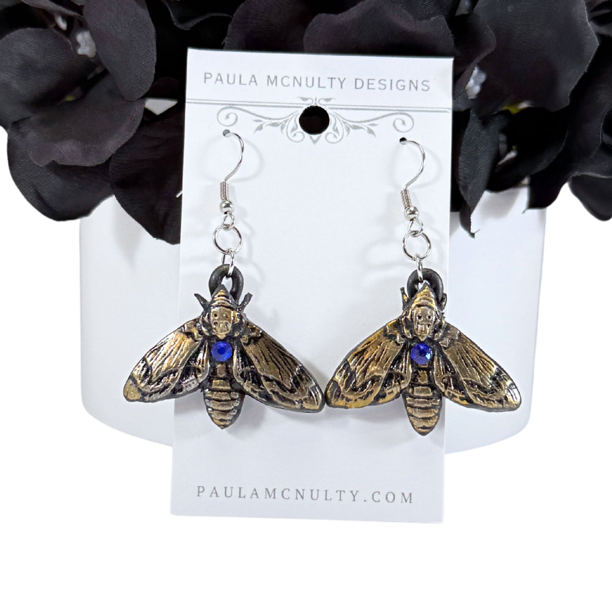 A pair of moth-shaped dangle earrings with gold-toned wings, dark metal bodies, and a small blue gemstone at the center, displayed on a white card labeled Paula McNulty Designs.