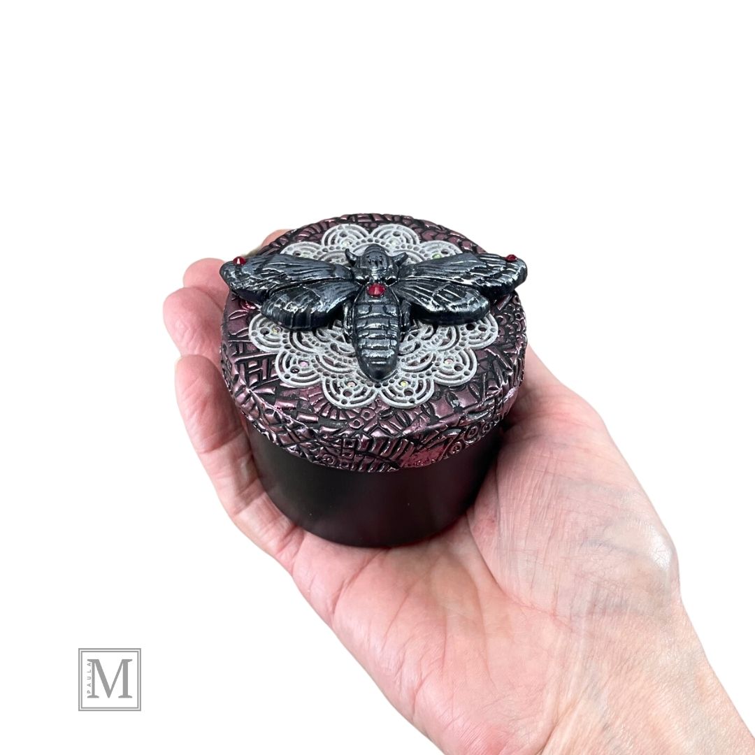 4oz black aluminum storage tin with handcrafted polymer clay purple textured lid with lace detail. with a hawkmoth with red crystal on abdomen. Held in model's hand