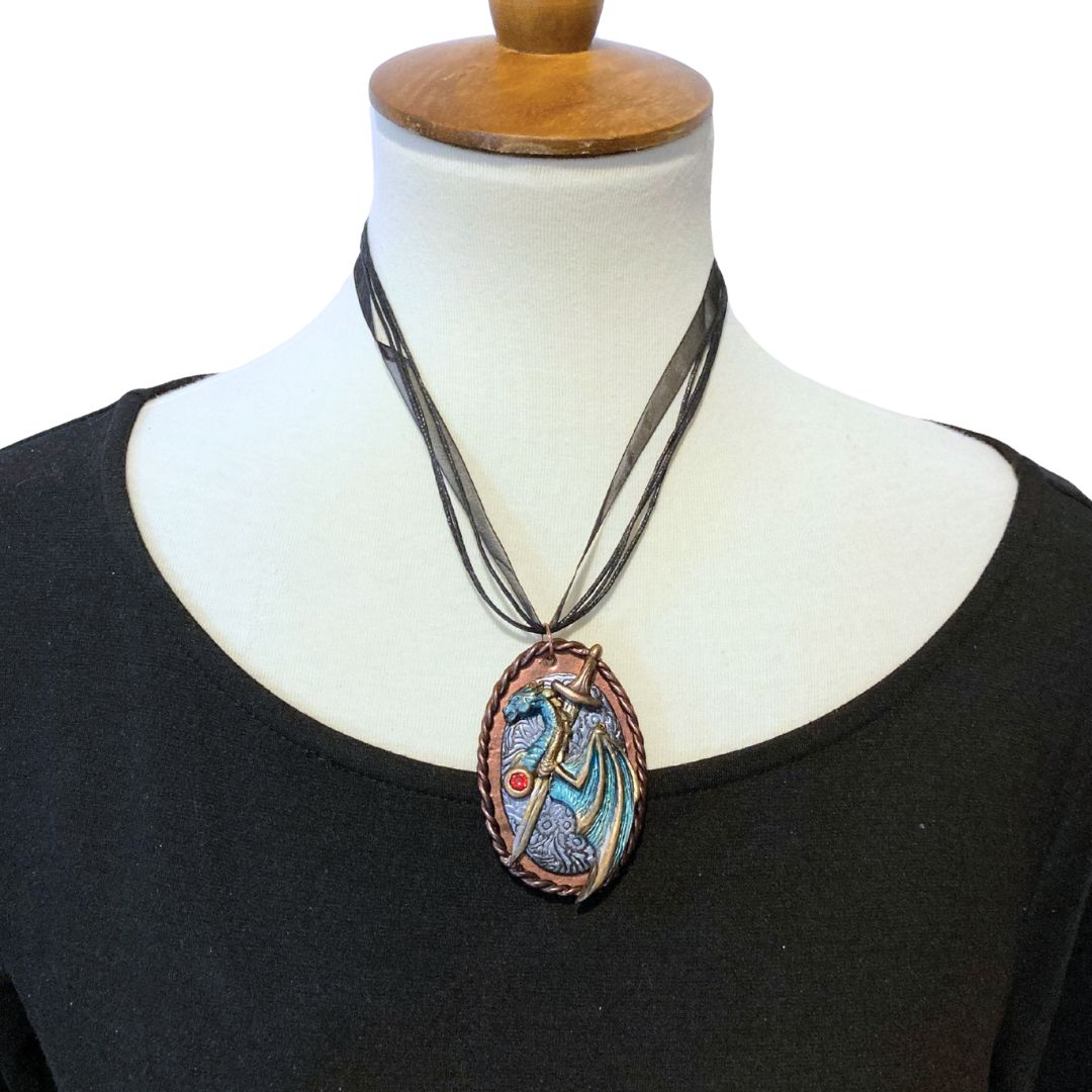 Dragon and sword necklace displayed on mannequin with black ribbon cord. High fantasy handmade jewelry inspired by dragons and epic adventures.