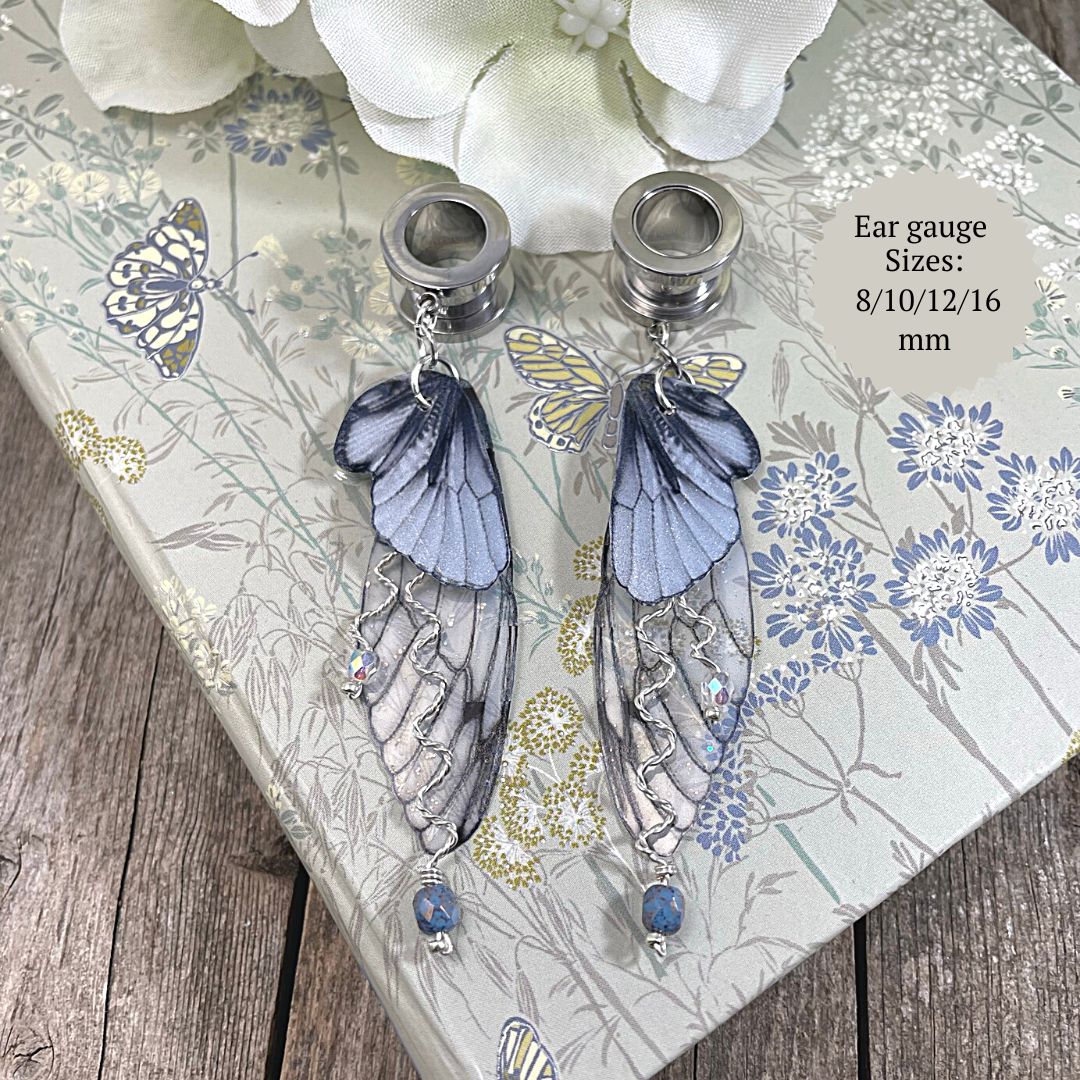 Double fairy wing earrings in silver with ear tunnels for stretched ears resting on a book with a flower in the background.