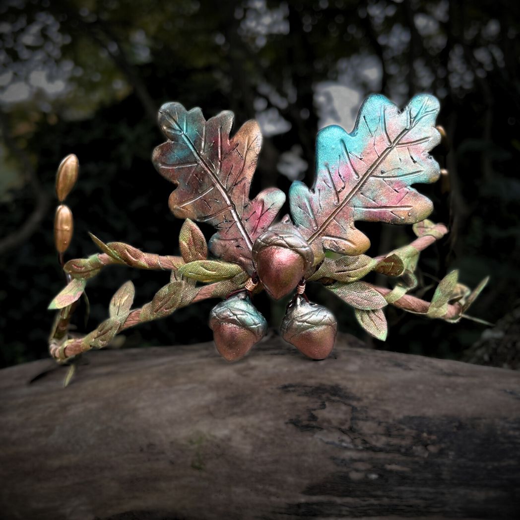 3 Acorns and 2 oak leaf crown in muted Fall colors with green and gold leaf vines and golden buds to the sides.  The crown in resting on a tree branch.