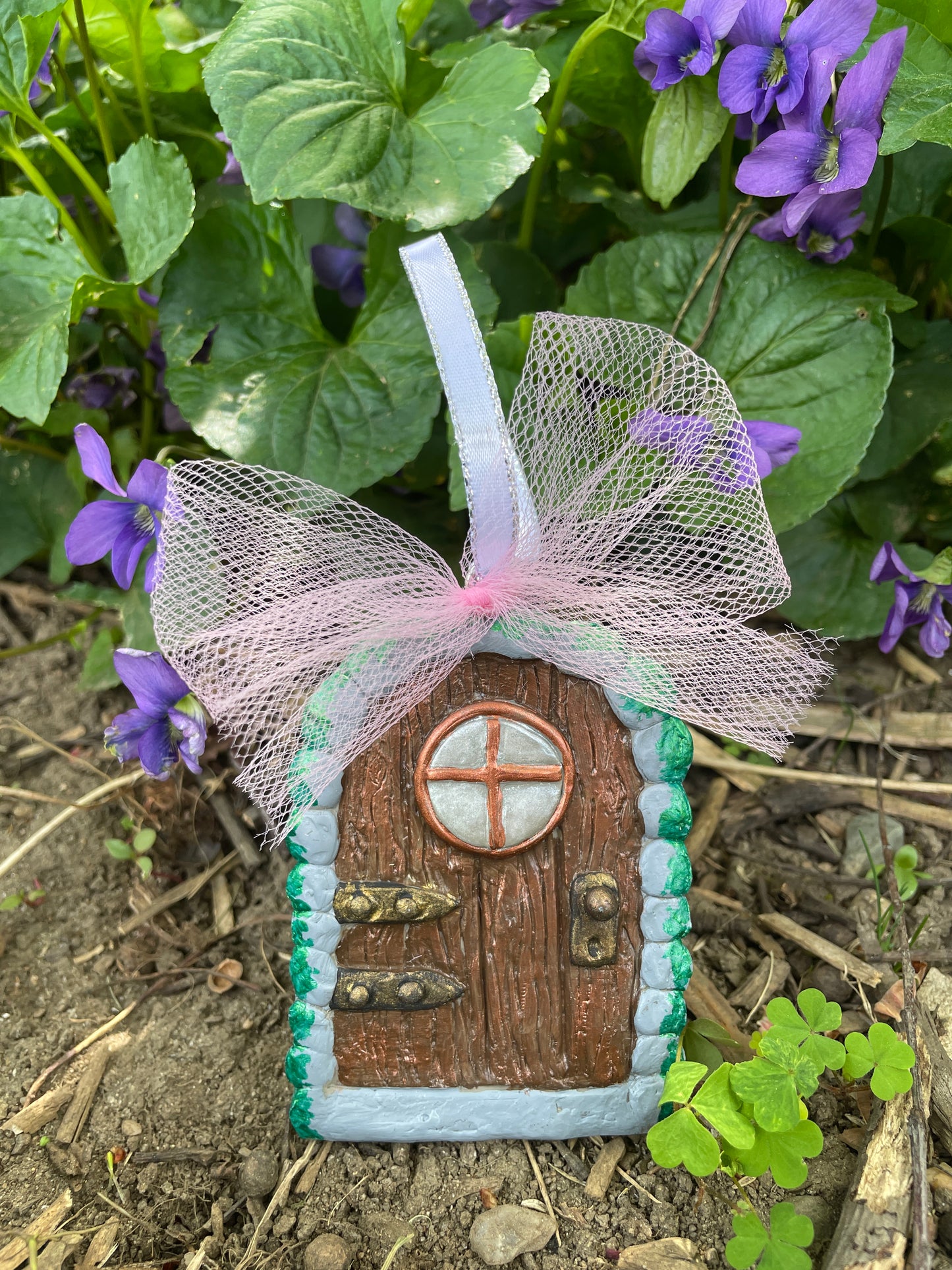 miniture arched elf door ornament with a with a pink tulle bow and ribbon hanger against green plants and purple flowers.