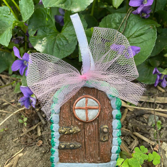 miniture arched elf door ornament with a with a pink tulle bow and ribbon hanger against green plants and purple flowers.