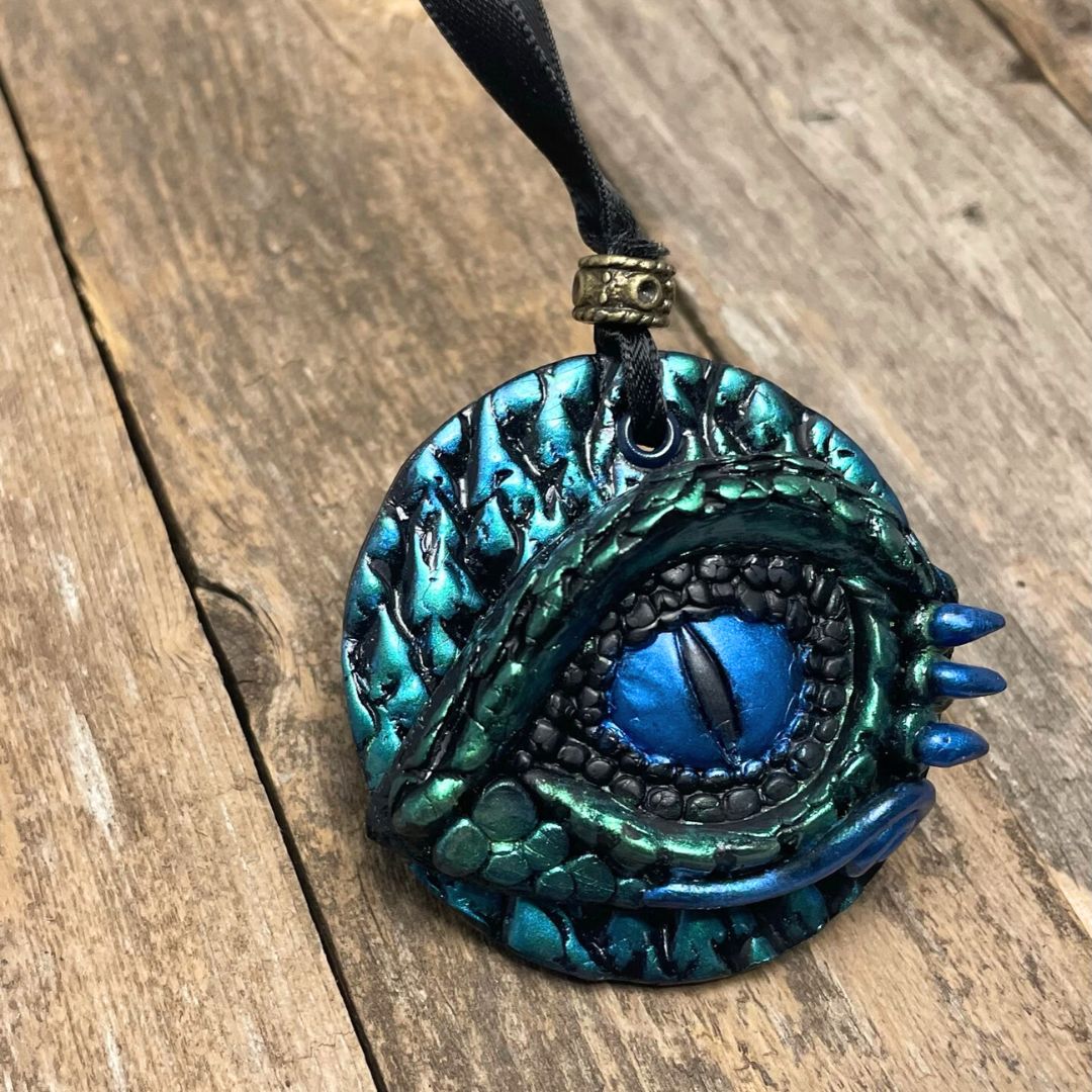 A round dragon eye ornament with metallic green and blue coloring with black satin ribbon held in a hand.