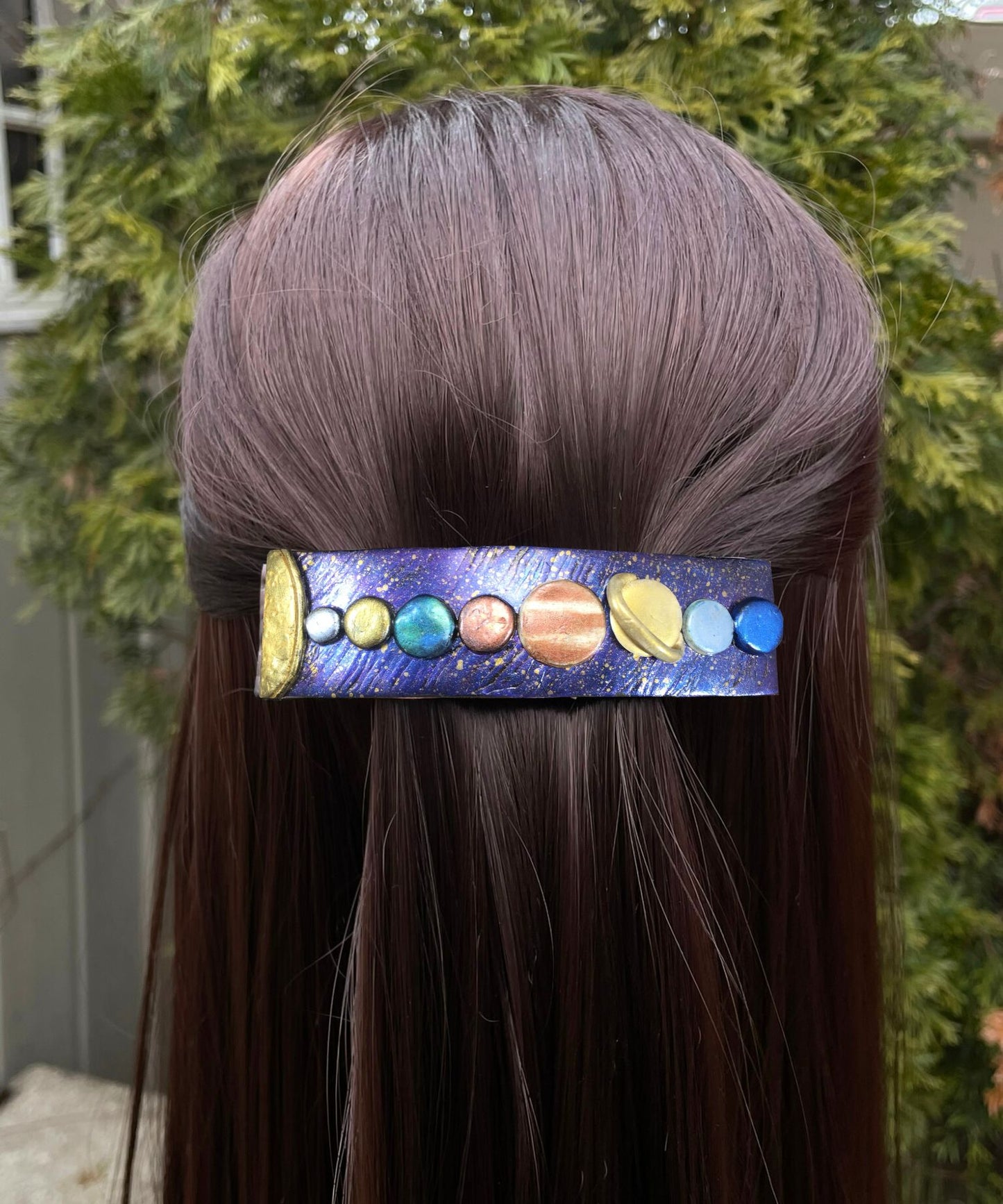 Handmade polymer clay hair barrette with purple/blue base with gold stars and hand painted solar system in clay represented in a line from the sun on far left  in model's hair.