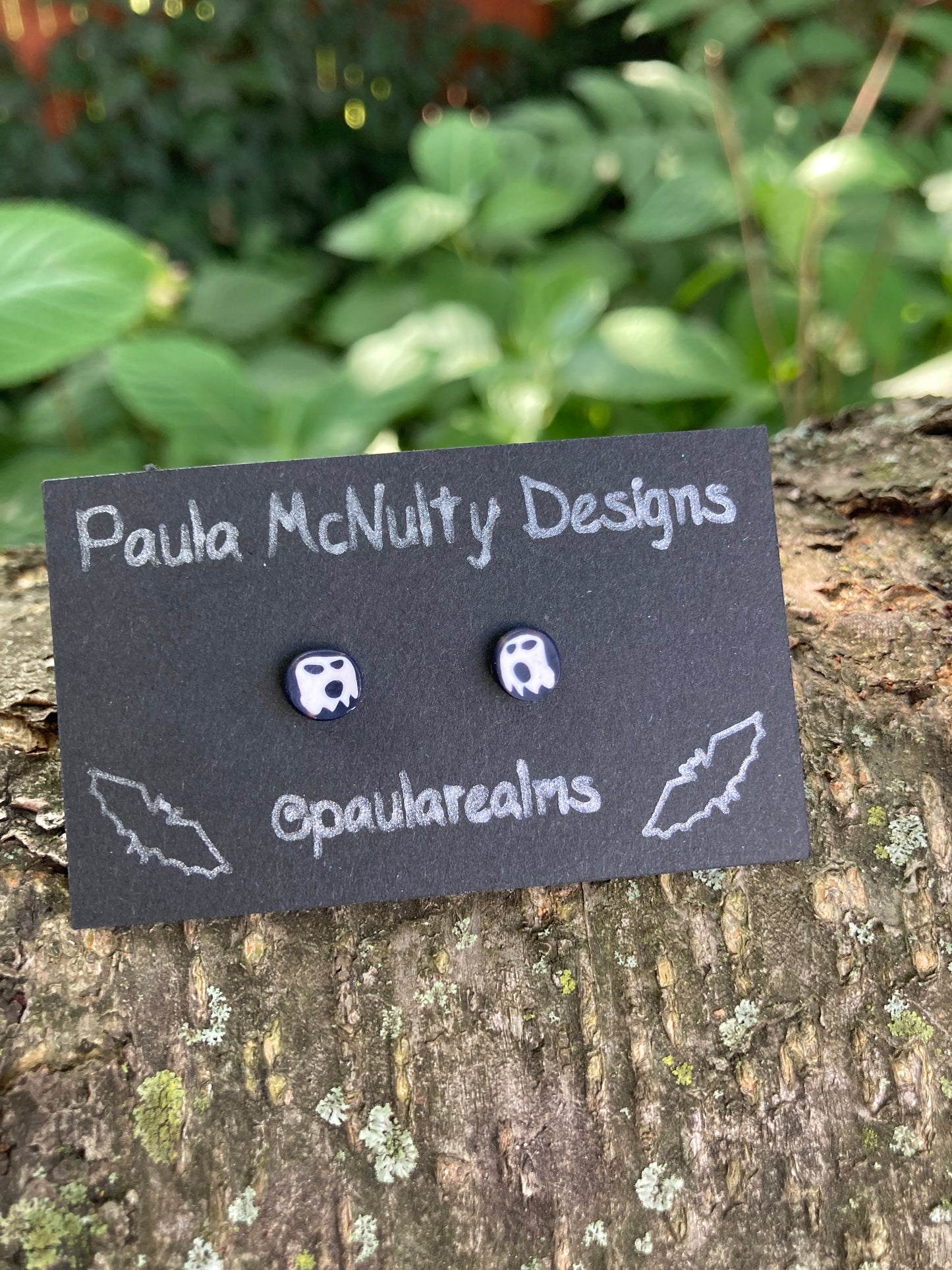 Tiny black round stud earrings with little white ghosts on a black earring card sitting on a tree branch.